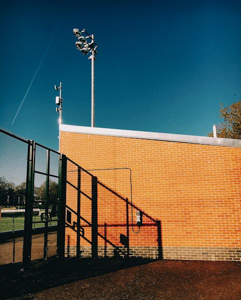Brick sports facility exterior with steel fencing and shadows, under a bright blue sky in Devon, England.